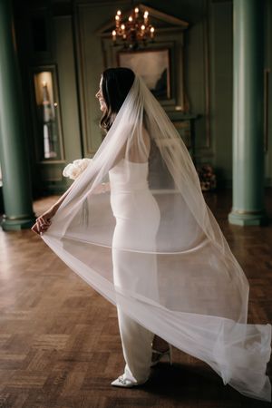 Elegant bride in fitted white dress twirls her flowing cathedral veil in a grand room with green columns and chandelier.