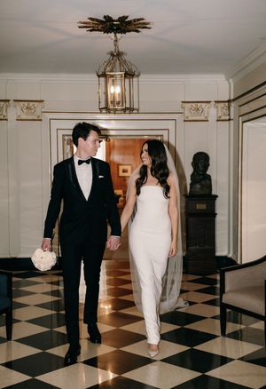 Elegant couple walks on black and white checkered floor beneath ornate vintage lantern in formal hall.