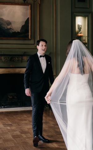 A bride in a flowing white veil and groom in black tuxedo share a moment in an elegant room with parquet flooring.