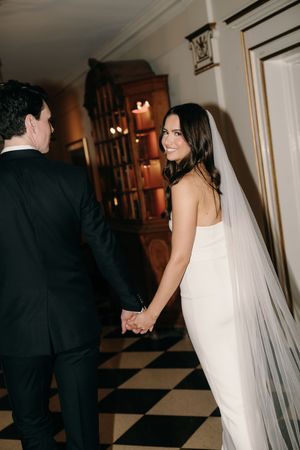 Newlywed couple holding hands while walking on a black and white checkered floor with a long white veil flowing behind.