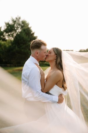 A bride in a white wedding dress with flowing veil kisses her groom in formal attire during an outdoor sunset photoshoot.