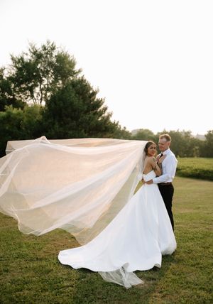 A romantic wedding couple embraces in a grassy field while the bride's long veil flows dramatically in the wind at sunset.