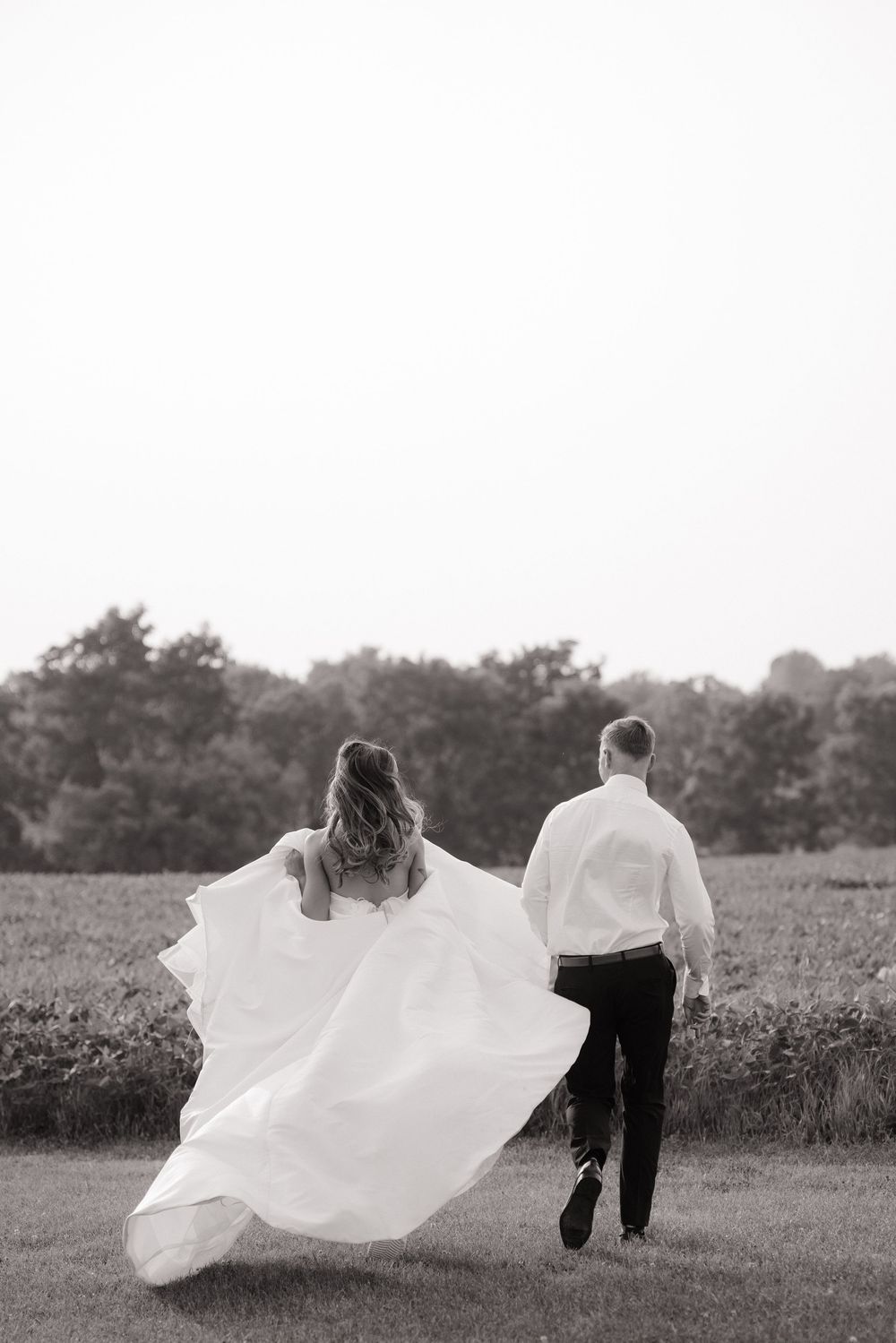 Black and white photo of couple walking in field as bride's white wedding dress flows elegantly in the breeze.