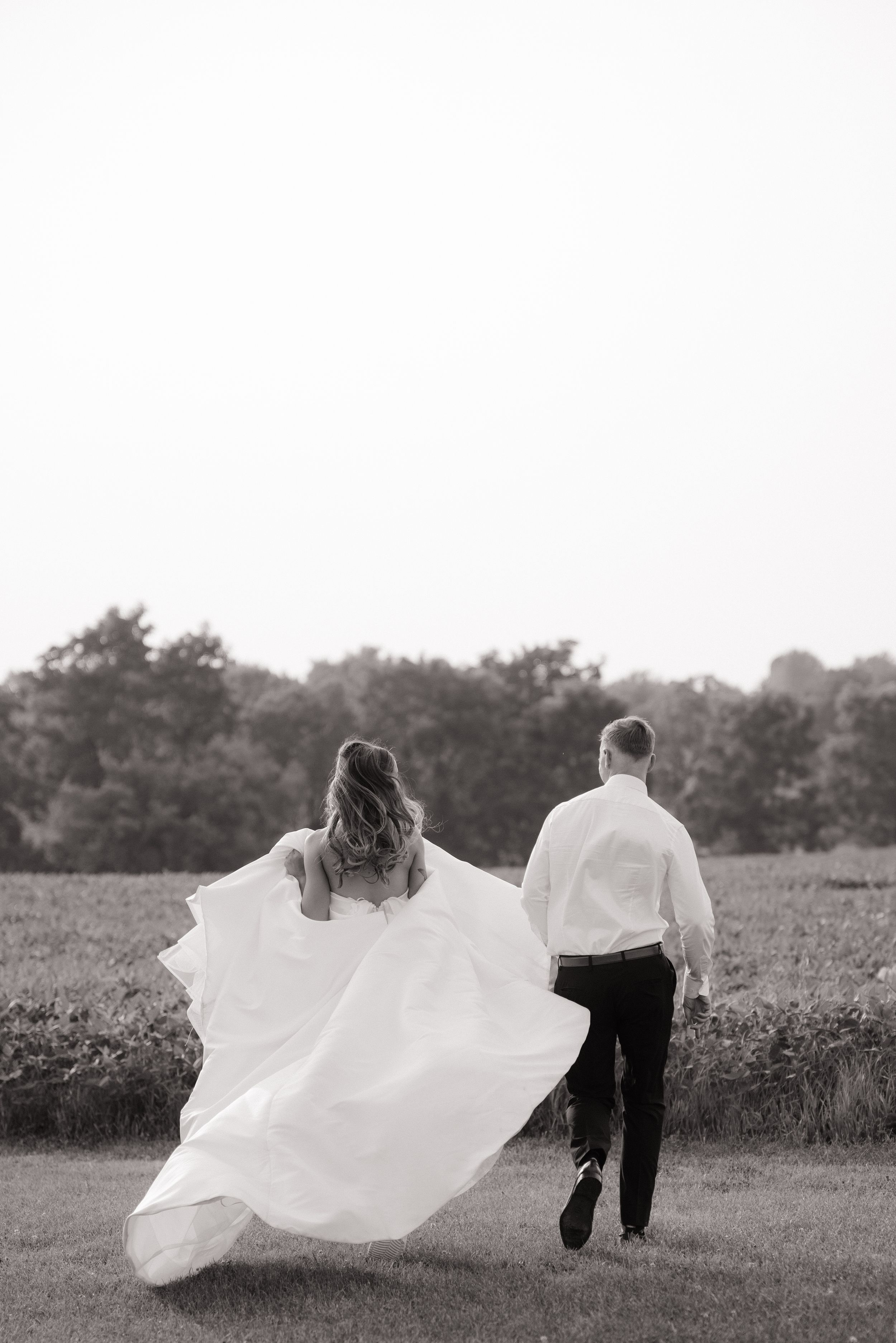 Black and white photo of couple walking in field as bride's white wedding dress flows elegantly in the breeze.
