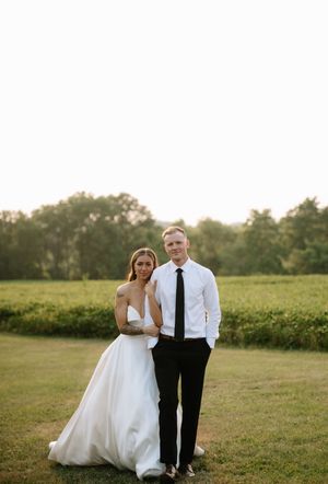 A couple poses together on their wedding day in a grassy field during golden hour light.