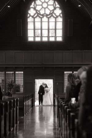 Black and white photo of bride and groom silhouetted in church doorway, with pews and stained glass window.