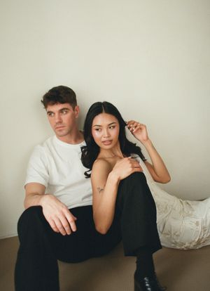 A man in a white t-shirt and woman in black sit together against a neutral wall in a minimalist fashion editorial.