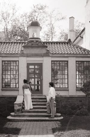 Black and white photo of a couple standing before a charming building with tile roof and large windows.