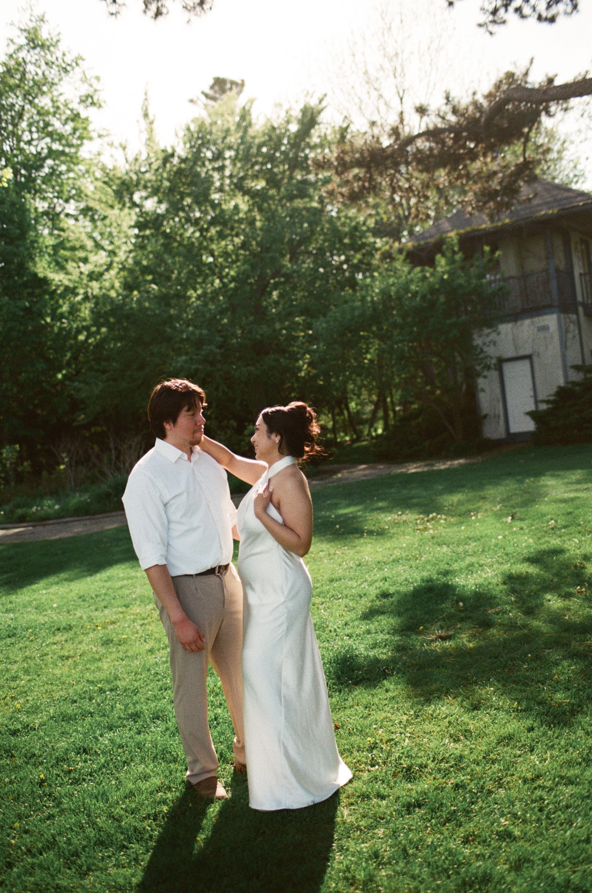 A couple in wedding attire share a romantic moment on a lush green lawn surrounded by trees.