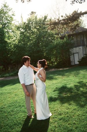 A couple in wedding attire share a romantic moment on a lush green lawn surrounded by trees.