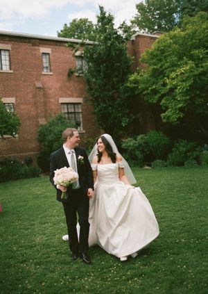 Bride and groom walking hand in hand on green lawn outside historic brick building on their wedding day.