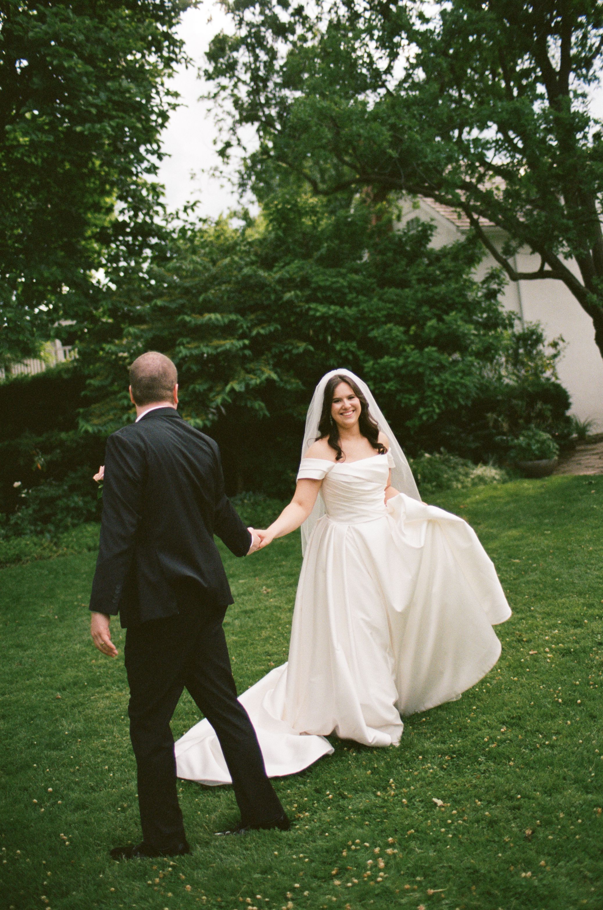 Smiling bride in white off-shoulder gown and veil holds hands with groom in black suit outdoors.