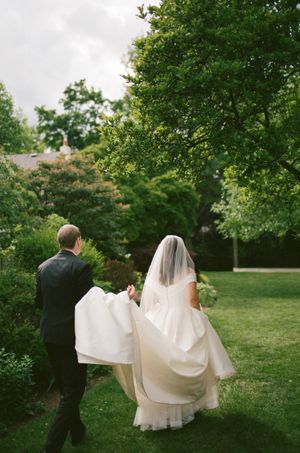 A bride and groom walk hand-in-hand through a lush green garden, her white gown and veil trailing behind them.