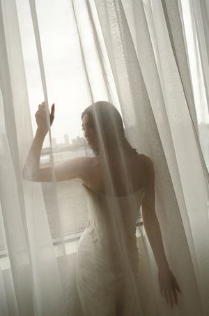 A bride in a white wedding dress stands behind sheer curtains by a window, bathed in soft natural light.