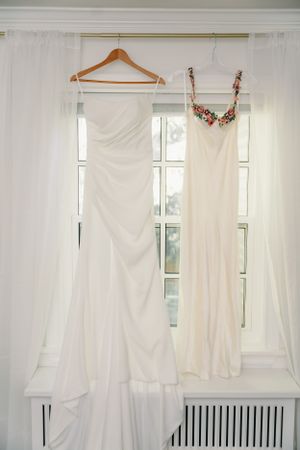 Two elegant white wedding gowns hanging on wooden hangers against sheer white curtains in front of a bright window.
