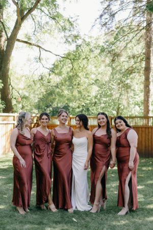 Wedding party in burgundy bridesmaid dresses standing with bride in white gown on grassy lawn with trees.