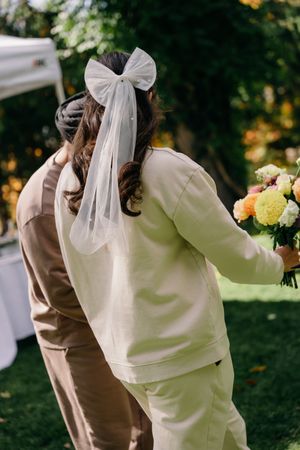 A person wearing a cream colored outfit with a white bow hair accessory leans forward in an outdoor garden setting.