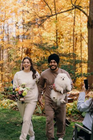Couple walking outdoors with their white Pomeranian dog against vibrant autumn foliage in a backyard setting.