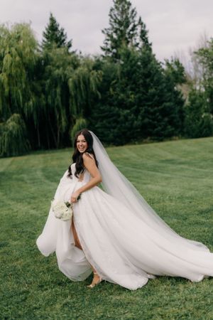 A bride in a flowing white wedding dress with a long veil poses on a green lawn with evergreen trees in the background.