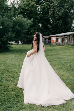 A bride in a flowing white gown and veil poses outdoors on a grassy lawn with trees and a rustic building in the background.