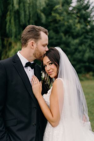 Romantic wedding couple embraces outdoors with willow trees in background during intimate portrait session.
