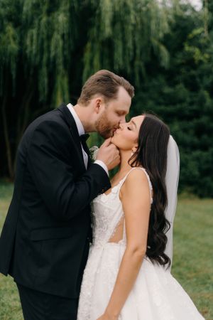 Bride and groom share a tender kiss in garden setting with dramatic willow trees behind them.