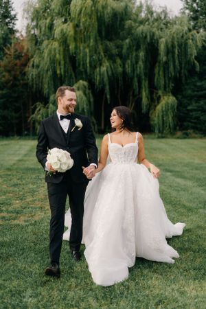 A wedding couple walks hand in hand on a green lawn with dramatic weeping willow trees in the background.