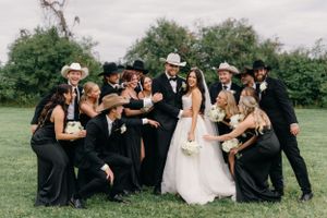 Wedding party in black and white attire poses playfully together on grassy field with bride in center.