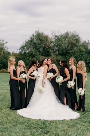A bridal party poses on a green lawn in matching black dresses holding white bouquets while celebrating a wedding.