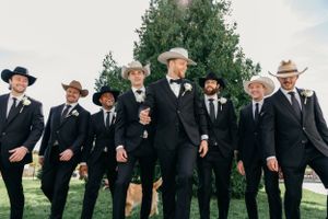 A group of groomsmen in black suits and hats pose together outdoors with trees in the background.