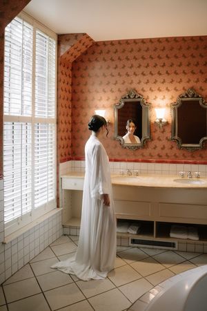 A white wedding dress hangs elegantly in a bathroom with floral wallpaper and shuttered windows.