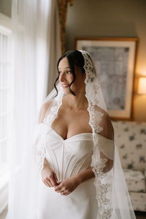 A bride wearing a delicate lace veil and strapless white wedding dress poses in soft natural light.