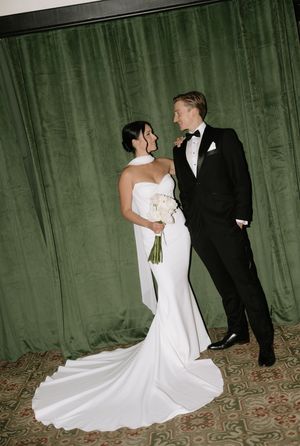A bride in a white mermaid gown and groom in black tuxedo share a moment against an emerald green curtain backdrop.