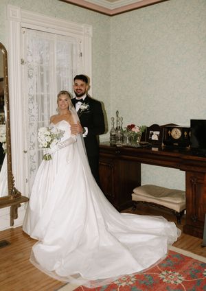 A bride in a flowing white wedding gown poses in an elegant room with hardwood floors and vintage furniture.