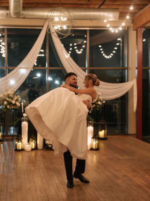 A romantic wedding couple dances in an industrial-chic venue with draped fabric and string lights creating an elegant atmosphere.