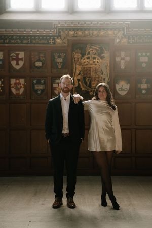Two people pose together in formal attire against a wood-paneled wall decorated with heraldic crests and shields.