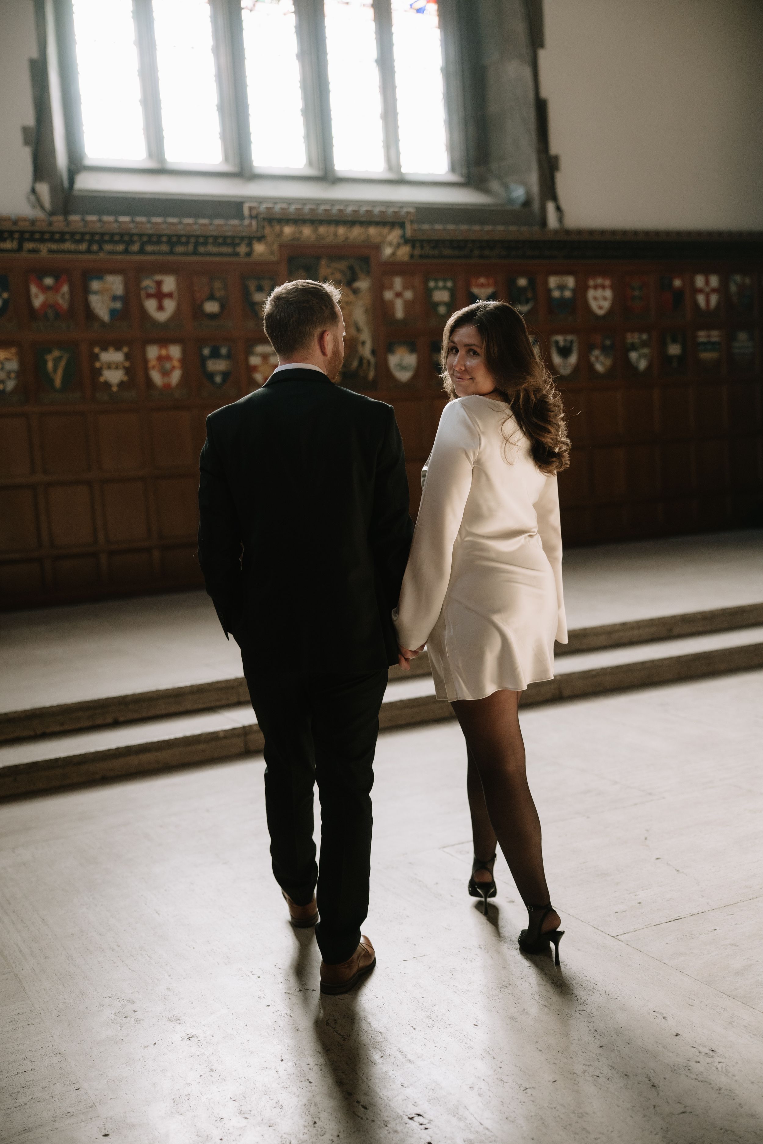 Two people walk together down a dimly lit hallway with ornate wall decorations visible in the background.