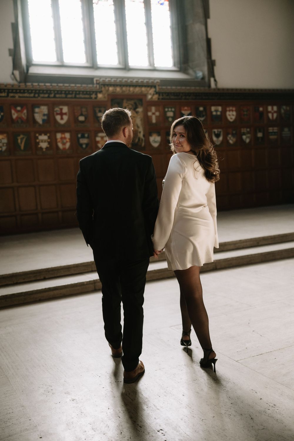 Two people walk together down a dimly lit hallway with ornate wall decorations visible in the background.