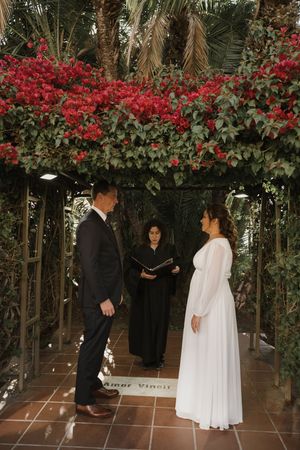 A wedding ceremony under an archway covered in vibrant red flowers and greenery.