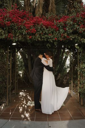 A couple embraces under an archway covered in red flowers during their wedding ceremony.