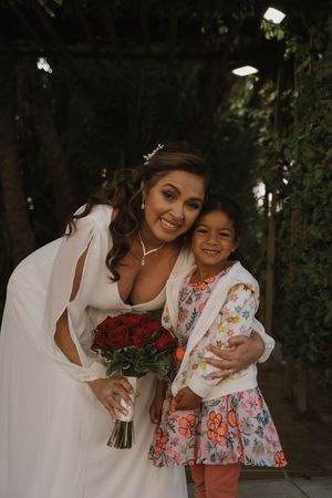 A bride in a white dress holds red roses while posing with a young girl in a floral dress outdoors.