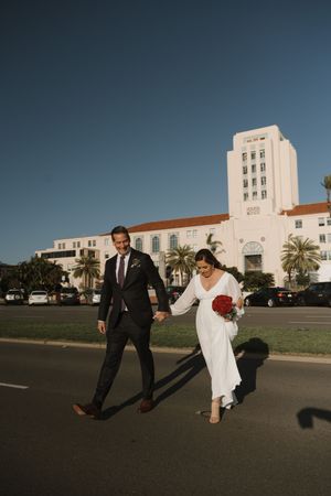 Newlyweds walk together in front of a historic white building with Spanish architecture.
