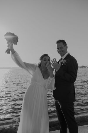 Couple celebrating on beach as bride raises bouquet triumphantly against ocean backdrop.