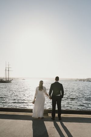 Silhouetted couple stands at waterfront looking out at sailboat on harbor at sunset.