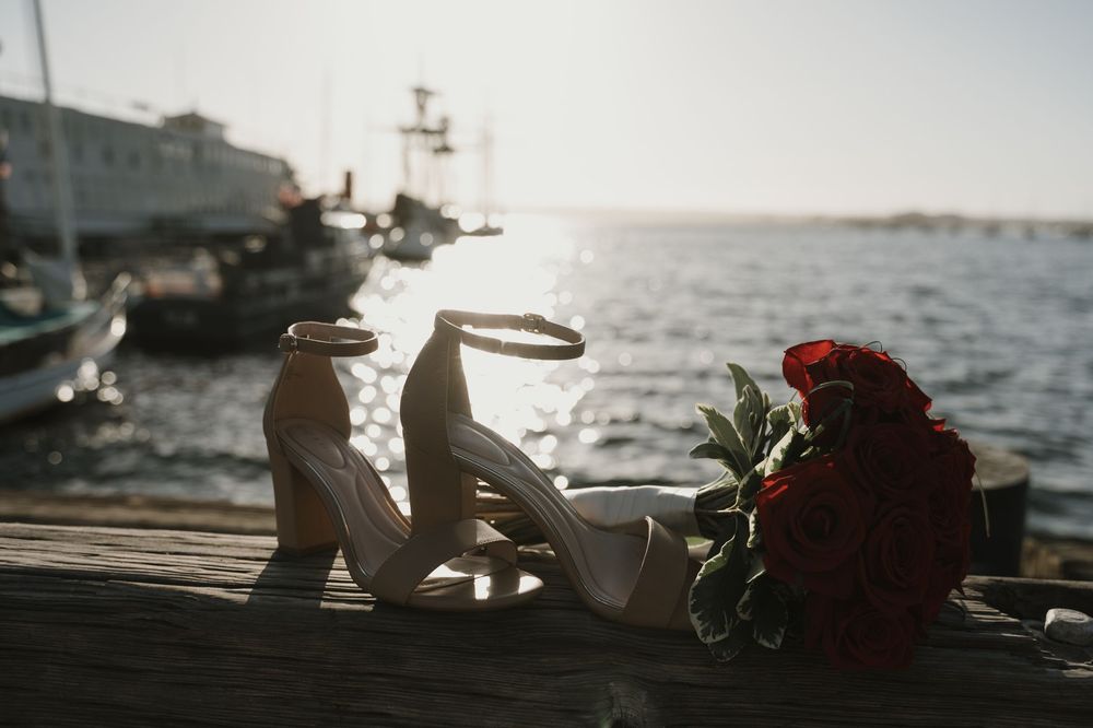 Wedding shoes and red rose bouquet arranged on wooden dock by the ocean.