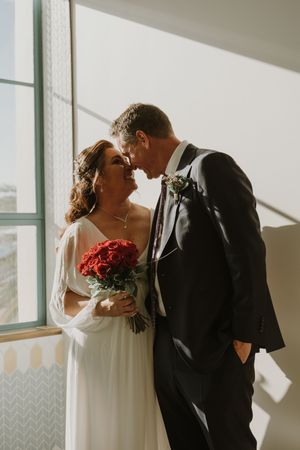 A couple shares an intimate moment by a window with red roses and sunlight streaming in.