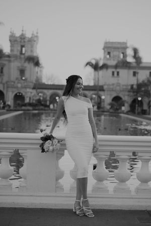 Black and white photo of person in wedding dress posing by ornate balustrade with church spires in background.