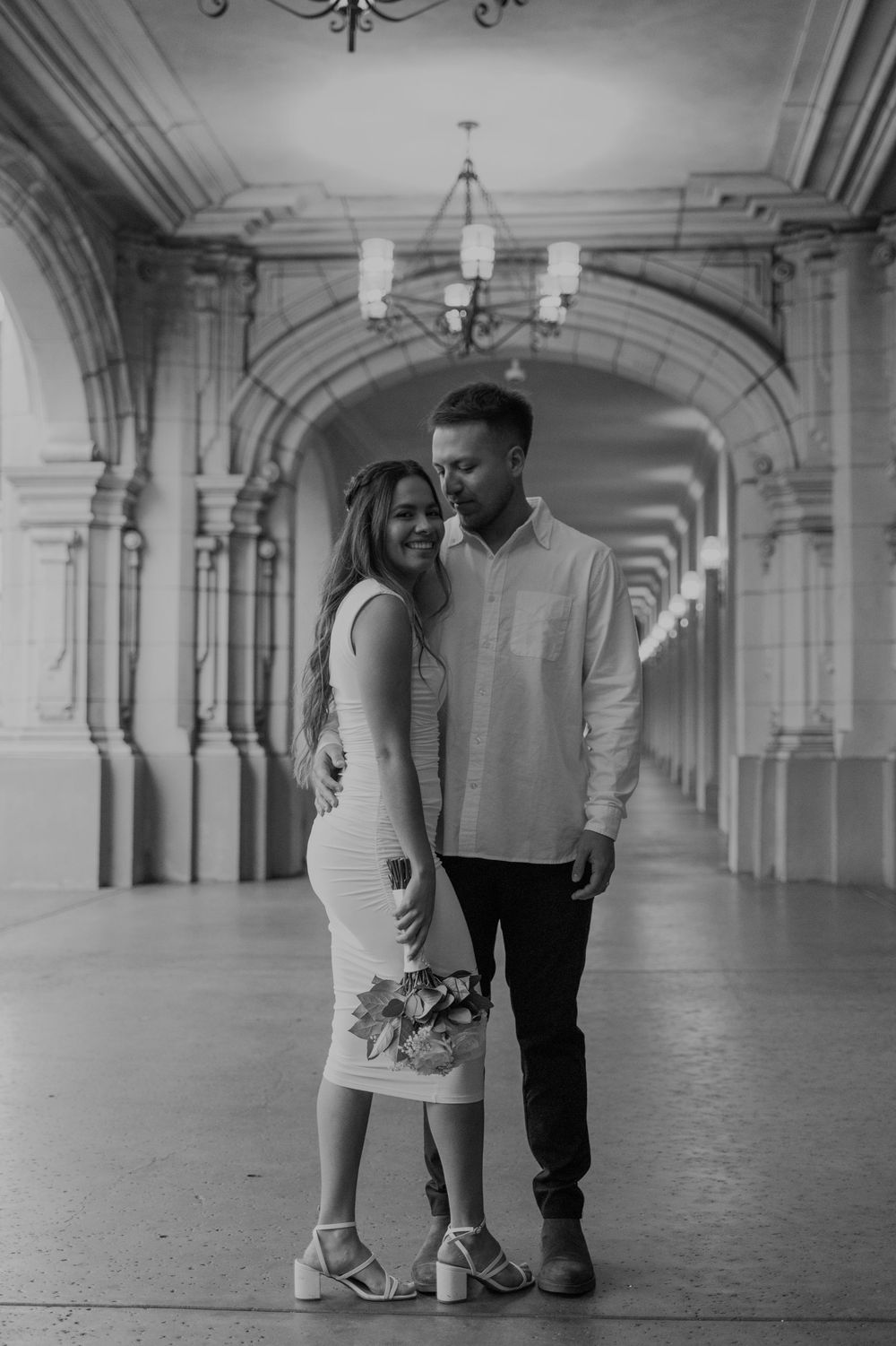 A couple poses romantically in an elegant archway corridor with a chandelier in black and white.