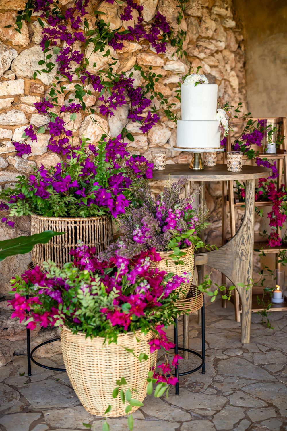 Elegant white wedding cake displayed on wooden table with vibrant purple bougainvillea flowers cascading down stone wall.