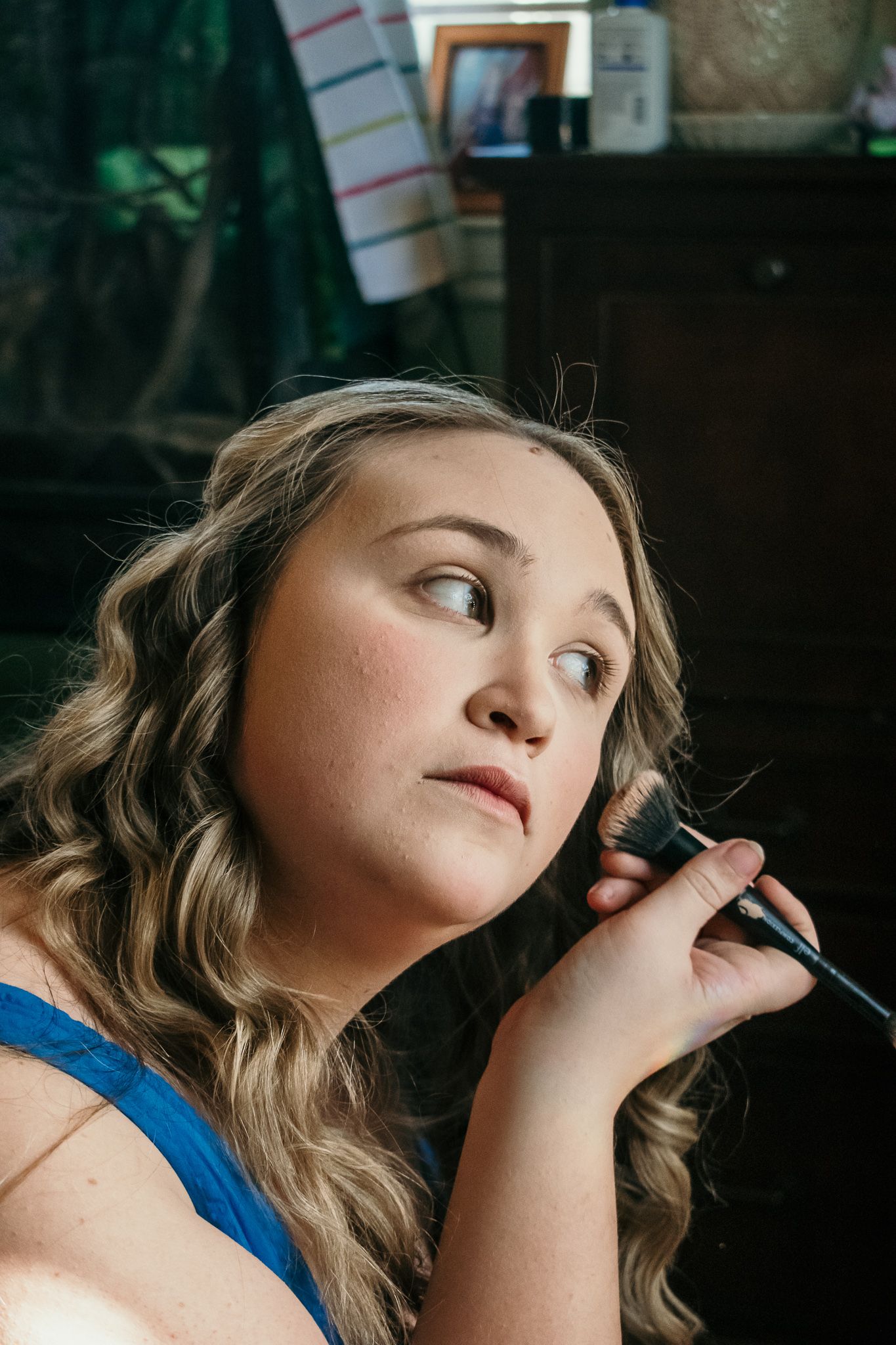 A person applies makeup with a brush while looking pensively to the side in a dimly lit room.